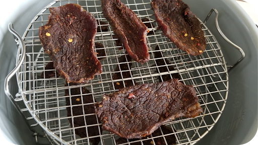 Thin strips of seasoned beef jerky on a wire rack inside an air fryer basket, showing the drying process in progress