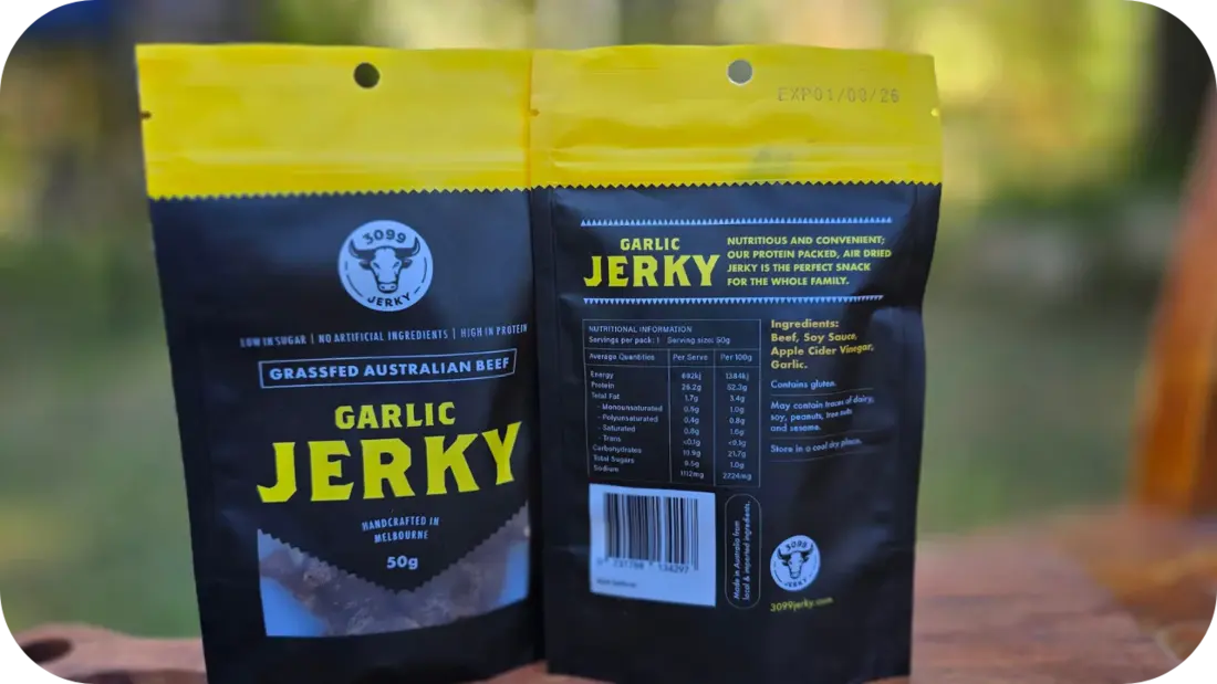 Two 3099 Garlic Jerky packs on a tray outdoors with pieces of jerky displayed in front, showing both front and back packaging.