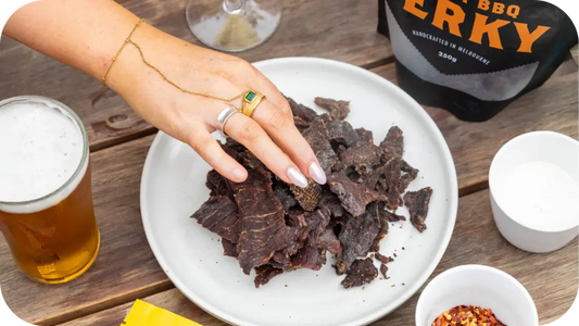 A hand picking jerky from a plate and a pack of 3099 smoky beside it with a glass of drink on a wooden table