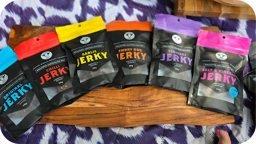 A row of 3099 Jerky packs in flavours like Original, Chilli, Garlic, Smoky BBQ, Teriyaki, and Salt & Vinegar on display.