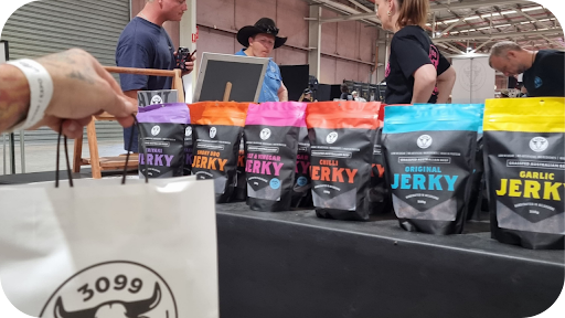 Colourful packs of 3099 Jerky displayed on a stall table with a shopper holding a branded bag in front.