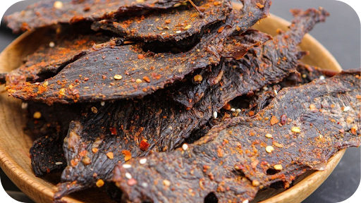 A wooden bowl filled with dark, textured pieces of beef jerky seasoned with visible red chili flakes and spices.