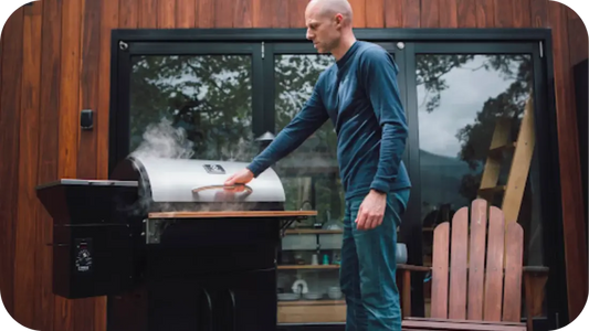 Man preparing beef jerky in a smoker outside a wooden cabin, adjusting temperature as smoke rises for authentic slow-cooked flavour.