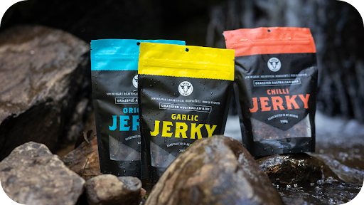 Three bags of 3099 Jerky in Original, Garlic, and Chilli flavors, resting on wet rocks in a flowing stream.