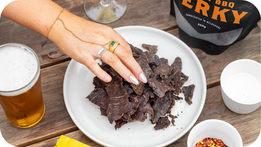 a hand picking jerky from a plate and a pack of 3099 smoky beside it with a glass of drink on a wooden table