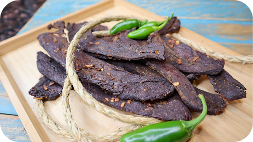 A wooden tray of spicy beef jerky strips garnished with green chilli peppers, showcasing a bold and flavourful snack.