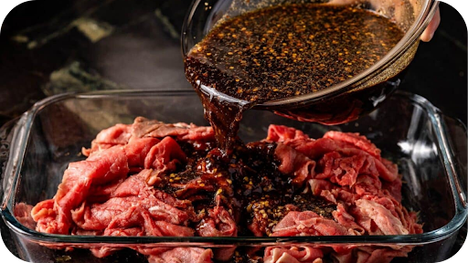A dark, seasoned marinade is poured from a glass bowl over a pile of thinly sliced raw beef in a glass baking dish.