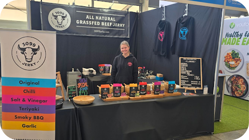 A smiling stallholder at a 3099 Jerky market stand showcasing grass-fed beef jerky packs, flavours, and branded merchandise.
