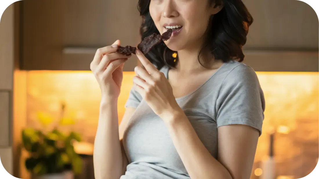 Pregnant woman in a grey t-shirt smiling softly while holding and eating a strip of beef jerky in a warm kitchen setting.