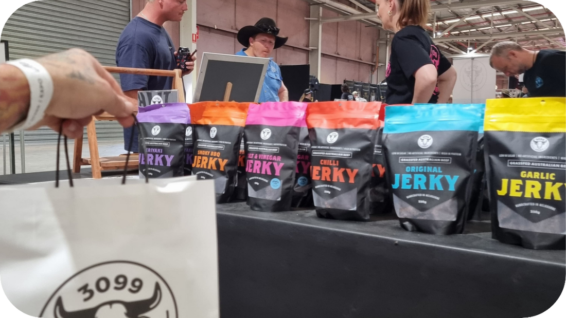 A market stall with colourful 3099 Jerky bags on display, including various flavours, while people browse, and a person holds a white shopping bag with the 3099 logo in the foreground.