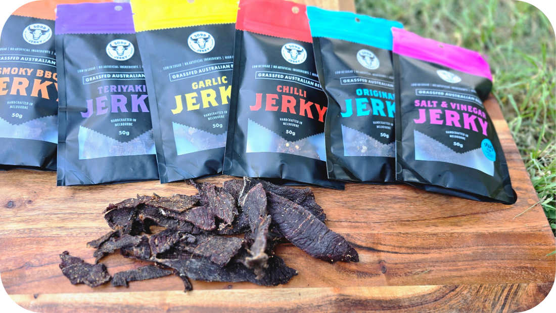 Variety of 3099 Jerky packs lined up on a wooden board with loose beef jerky pieces in front, showcasing different flavours.