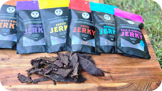 Variety of 3099 Jerky packs lined up on a wooden board with loose beef jerky pieces in front, showcasing different flavours.
