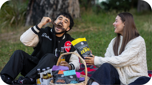 A young man and woman enjoying garlic beef jerky at a picnic, with drinks and snacks in a basket.
