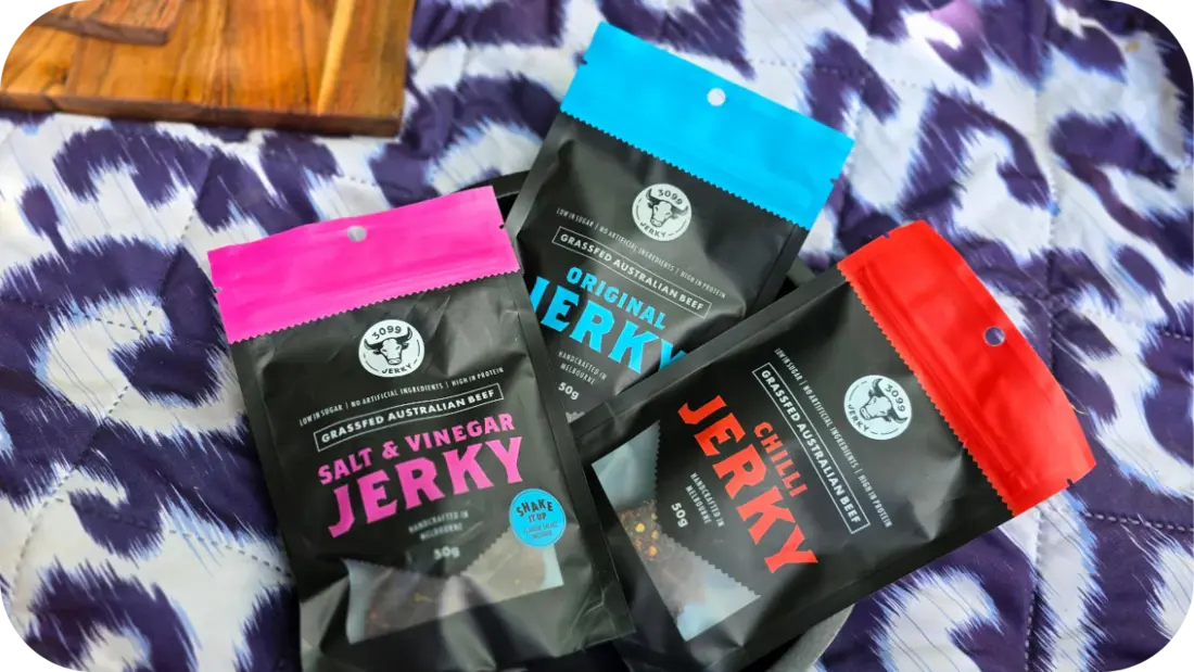 Three bags of 3099 Jerky in Salt &amp; Vinegar, Original, and Chilli flavors, fanned out on a purple and white animal-print blanket.