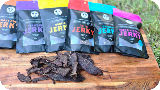 Variety of 3099 Jerky packs in flavours like Teriyaki, Garlic, Chilli, and Salt & Vinegar displayed with jerky pieces on wood.