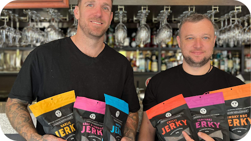 Two smiling men holding assorted 3099 Jerky wholesale packs inside a café bar, showcasing premium beef jerky supply for retailers.